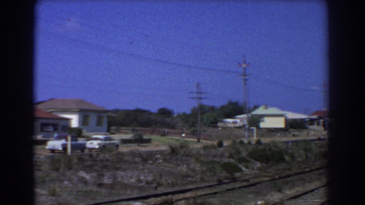 1967 Perth suburban houses seen from a moving train, with red-tiled roofs, parked cars, rail-side scrub, and power lines.