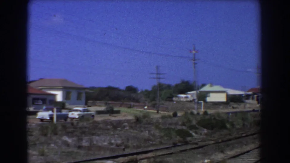 1967 Perth suburban houses seen from a moving train, with red-tiled roofs, parked cars, rail-side scrub, and power lines.