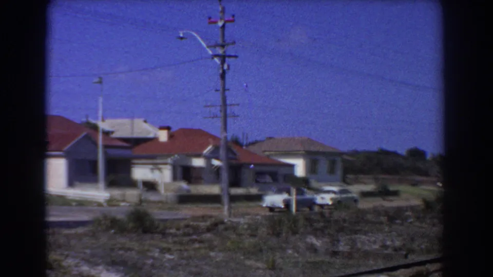 1967 Perth residential street view from a train, showing single-story homes, red tile roofs, parked cars, poles, and scrubby foreground.