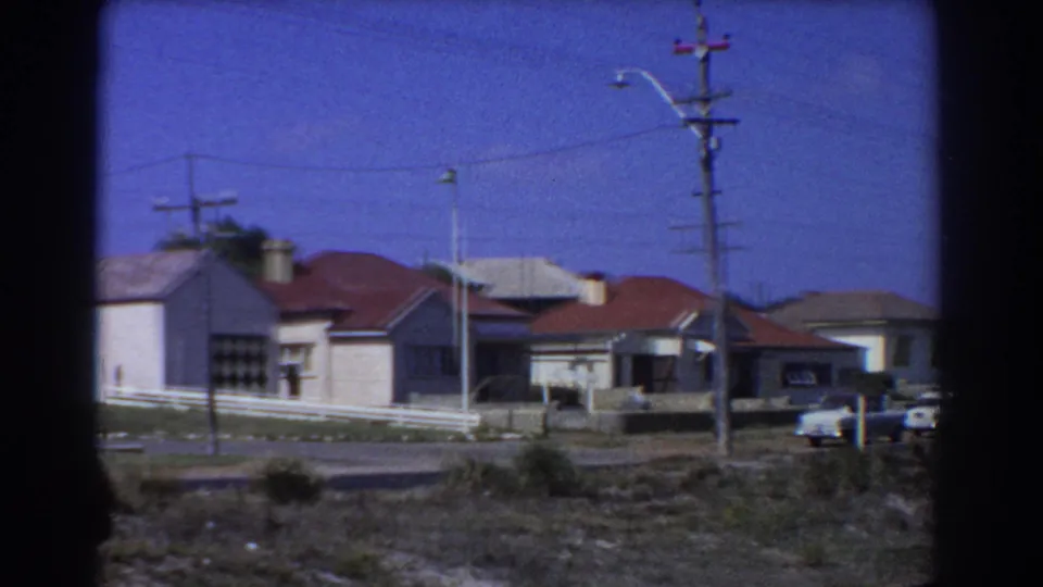 Late-1960s Perth suburban houses pass by the train, with red roofs, parked cars, rail-side scrub, poles, and overhead wires.