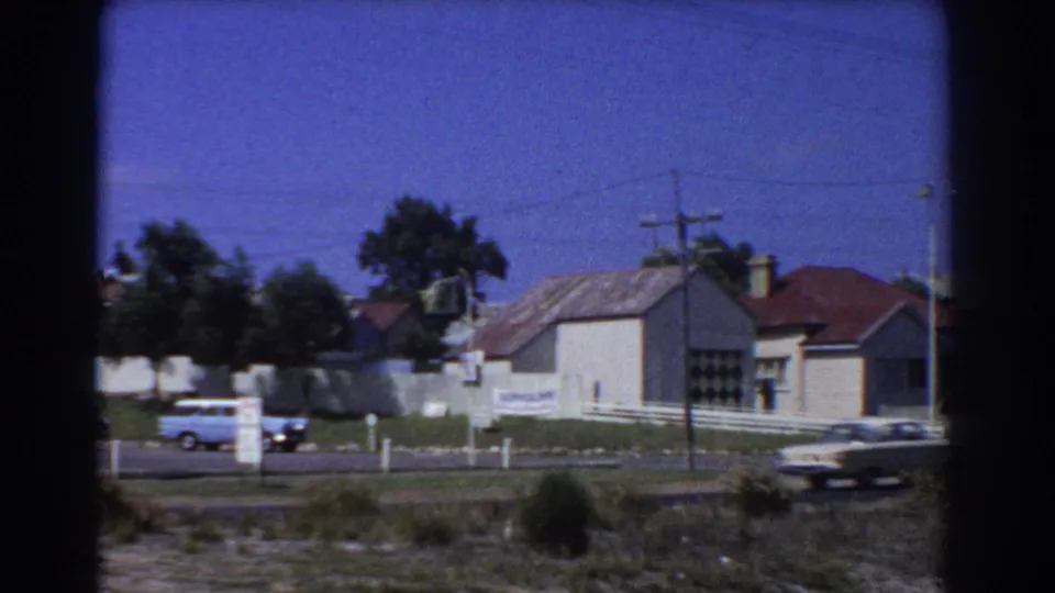 1967 Perth street scene with classic cars moving past low houses, fences, and utility wires under a blue sky.