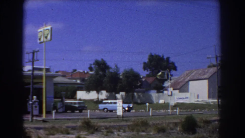 1967 Perth roadside view with a gas-station sign, passing cars, low houses, and visible film gate edges.