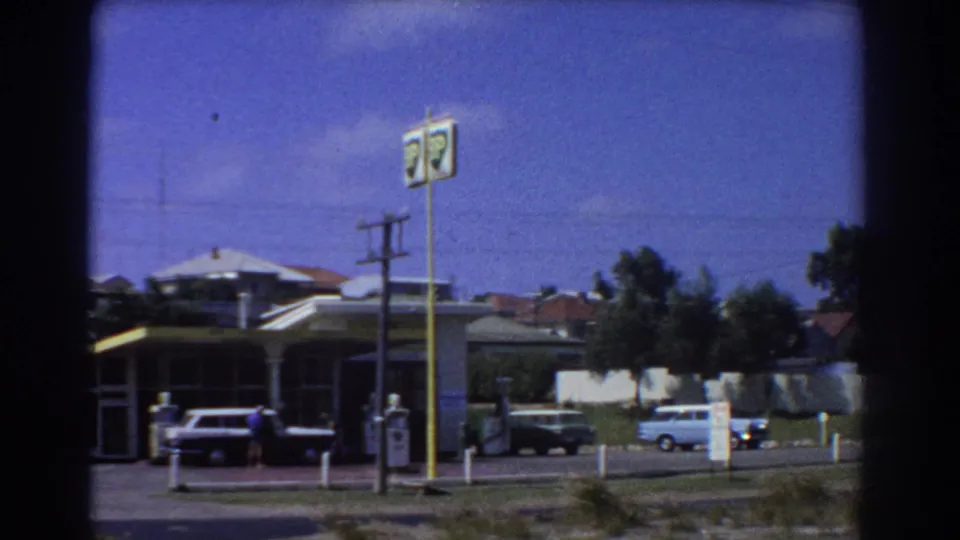 1967 Perth gas-station forecourt with classic cars, pump area, tall signs, and suburban houses behind it.