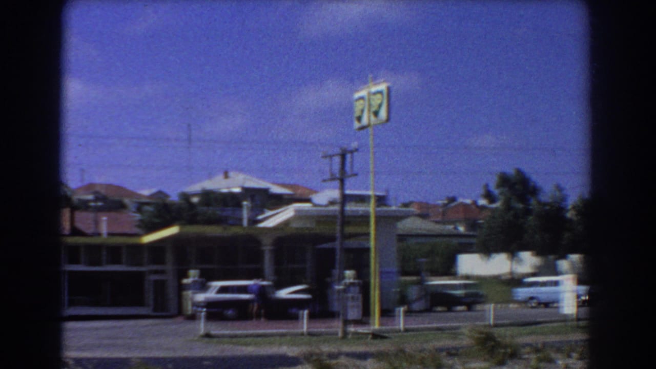 Service station forecourt with vintage cars, pumps, tall signpost and houses behind.