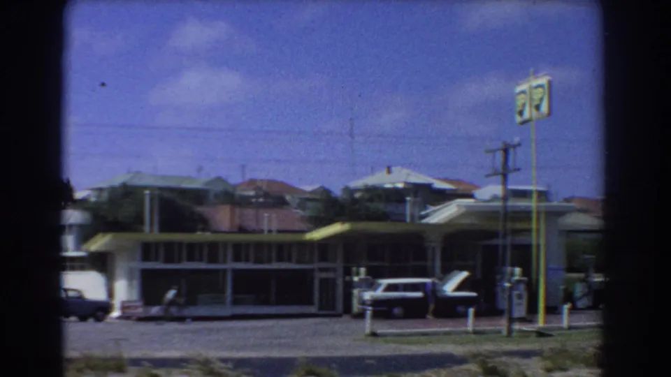 1967 Perth frame with a low service-station building, cars at the pumps, a raised hood and tall roadside signage.