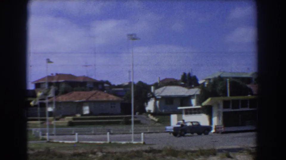 1967 Perth frame with a dark car near the service station frontage, tall street poles, suburban houses and blue sky with clouds.