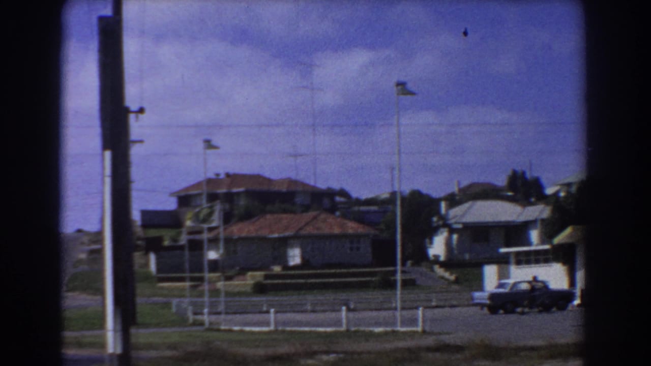 1967 Perth suburban street with red-roofed houses, a parked sedan, low fences, and utility poles under a cloudy blue sky.