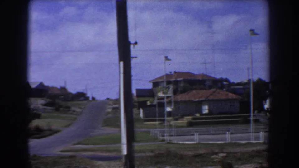 Perth suburban road in 1967, seen from a home-movie camera with houses, fences, poles, and open sky.