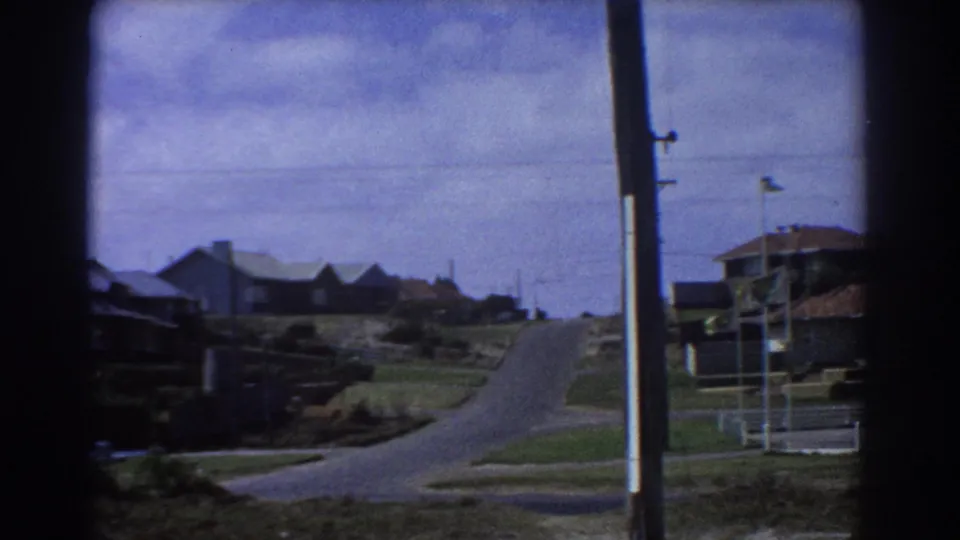 Home-movie view of a quiet 1967 Perth residential street with a curving road and low suburban houses.