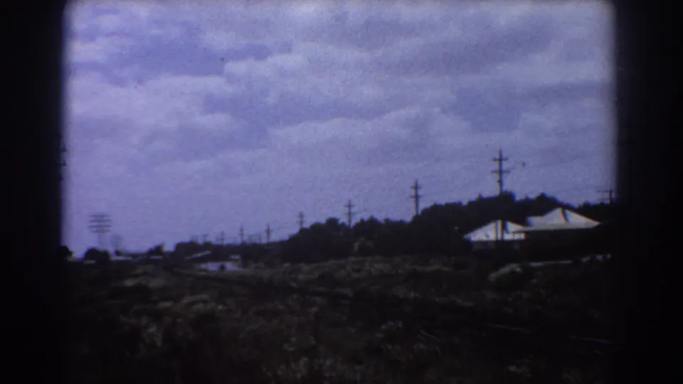 Moving 8mm view near Perth in 1967 with low white-roofed buildings, utility poles, trees, and rough grass beside the rail corridor.