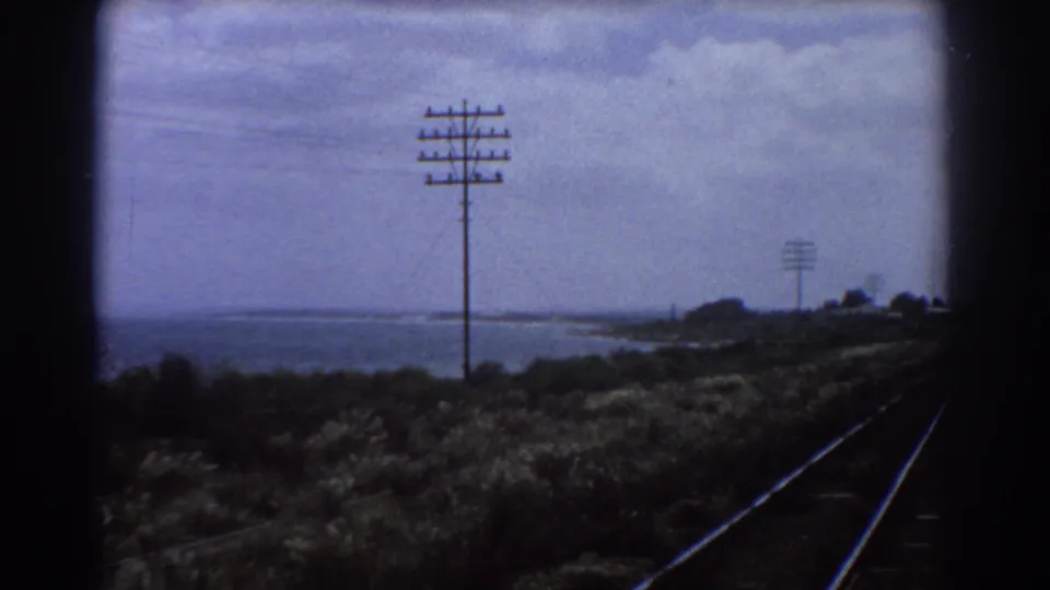 1967 Perth train-window view showing rails, tall utility poles, scrubby embankment, and a body of water under a cloudy sky.