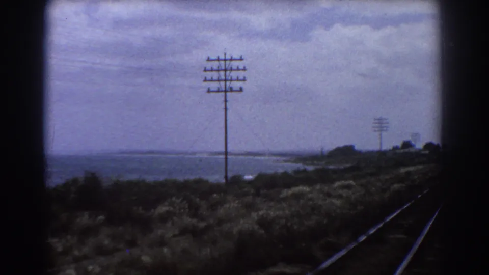 Perth-area 1967 rail landscape with tracks at right, utility poles, low vegetation, and water visible beyond the embankment.