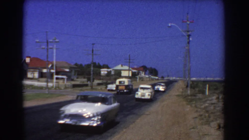 1967 Perth suburban street scene with mid-century cars traveling past modest houses, utility poles, and a dry roadside edge.