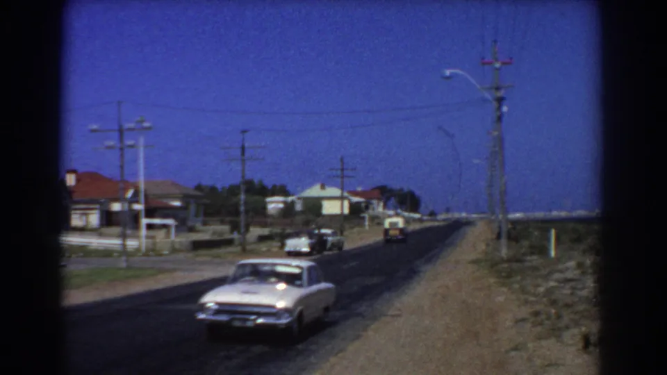 1967 Perth 8mm frame showing a light-colored car approaching on a suburban road beside houses, utility poles, and dry verge.