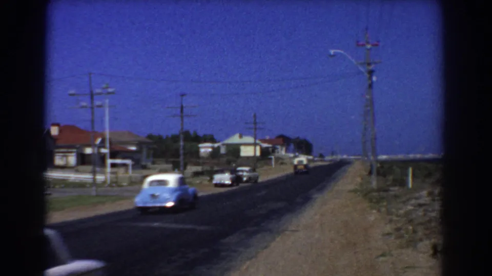 1967 Perth-area suburban road scene with classic cars passing low houses, utility poles, overhead wires, and a dirt roadside shoulder.