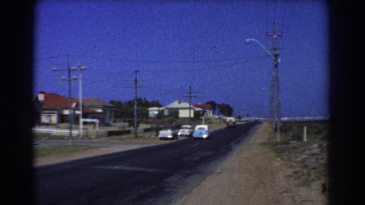 Cars travel along a paved road beside houses, poles, and open roadside ground.