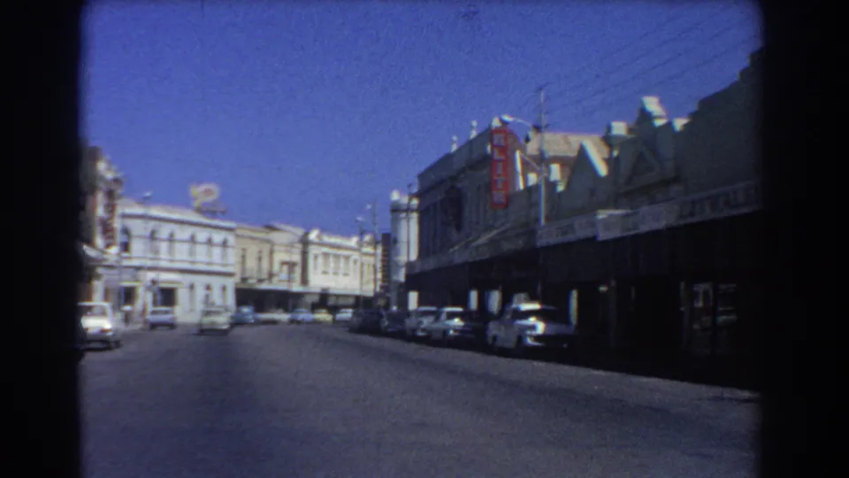 1967 Perth commercial street view showing awning-front buildings, parked classic vehicles, overhead lines, and a lightly trafficked roadway.