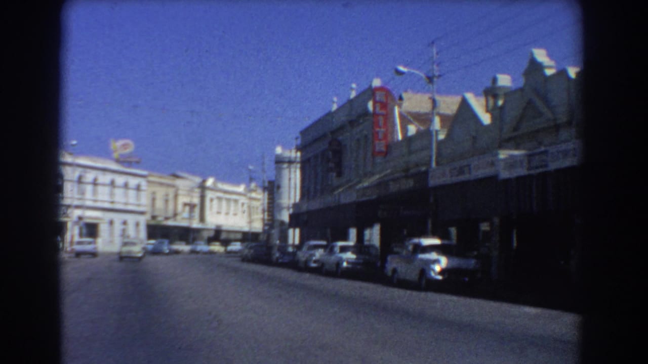 1967 Perth commercial street view with parked mid-century cars, shopfront awnings, and bright sun from a moving 8mm camera.