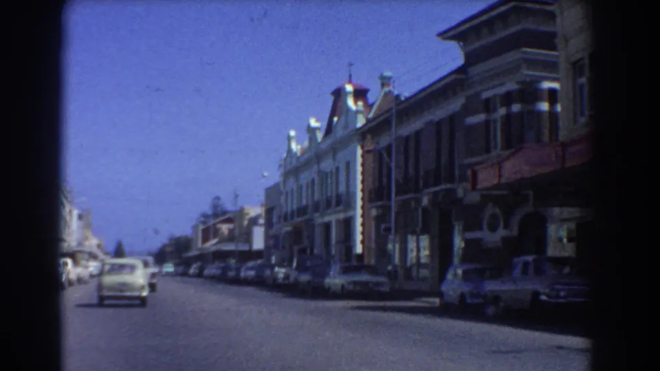 From a moving vehicle, the 8mm frame shows a car ahead on a Perth commercial street with rows of parked vehicles and older facades.