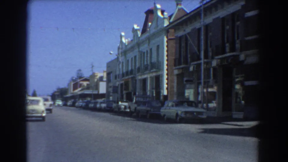 Late-1960s Perth street footage shows a commercial block, curbside parked cars, and traffic filmed from a moving vehicle.