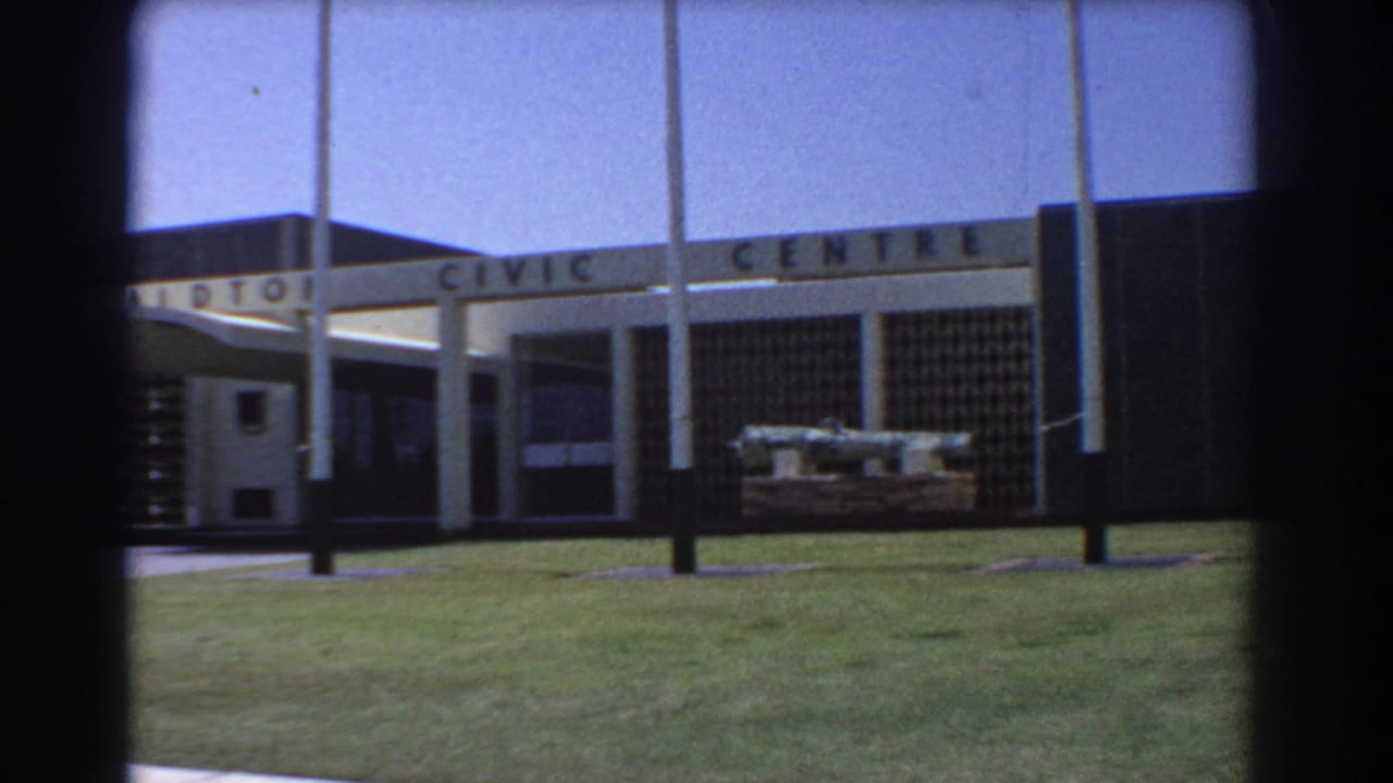 Geraldton Civic Centre exterior with lawn, flagpoles, walkway, and dark 8mm frame edges.