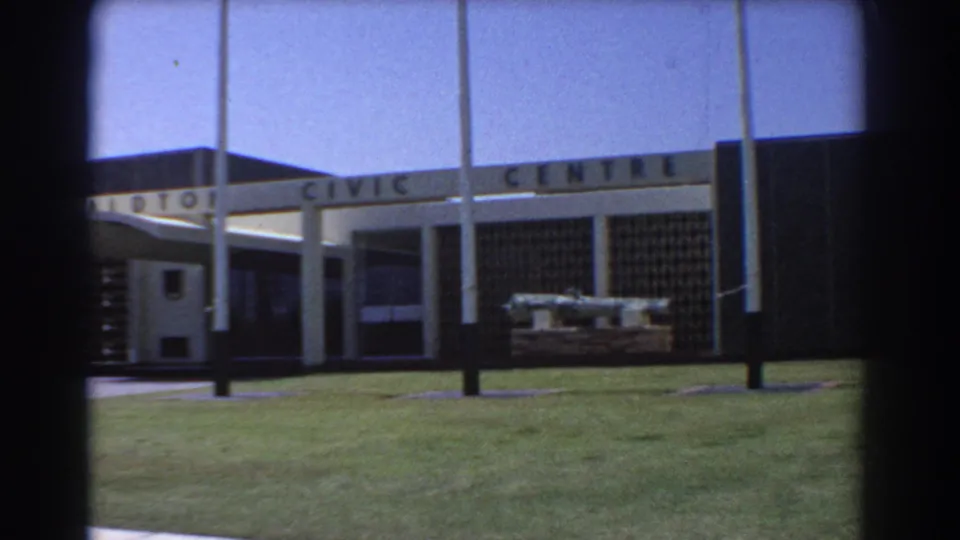 1967 8mm view of the Geraldton Civic Centre exterior in Australia, showing the facade, mowed lawn, flagpoles, and walkway.