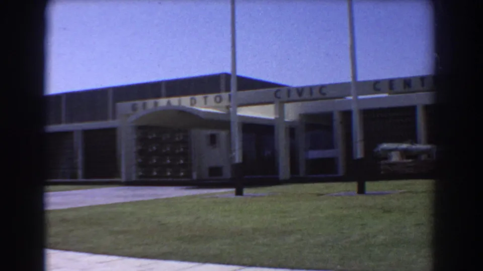 1967 8mm frame of the Geraldton Civic Centre entrance, with an arched canopy, flagpoles, trimmed lawn, and clear sky.