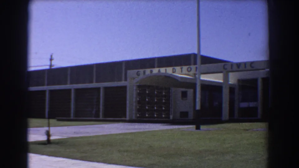 1967 8mm frame showing the Geraldton Civic Centre facade in Australia, with entrance canopy, flagpoles, lawn, walkway, and blue sky.