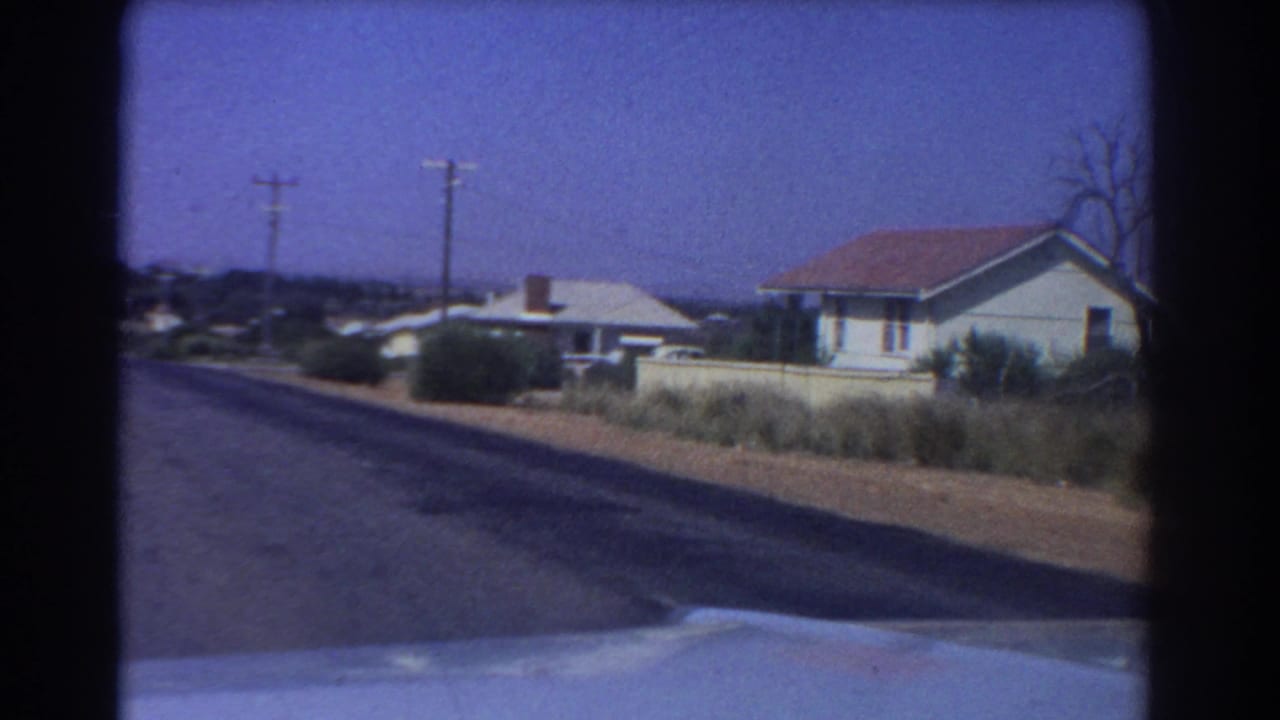 1967 Perth home-movie view along a suburban road, with a car hood at bottom, tiled-roof houses, low shrubs, and utility poles.