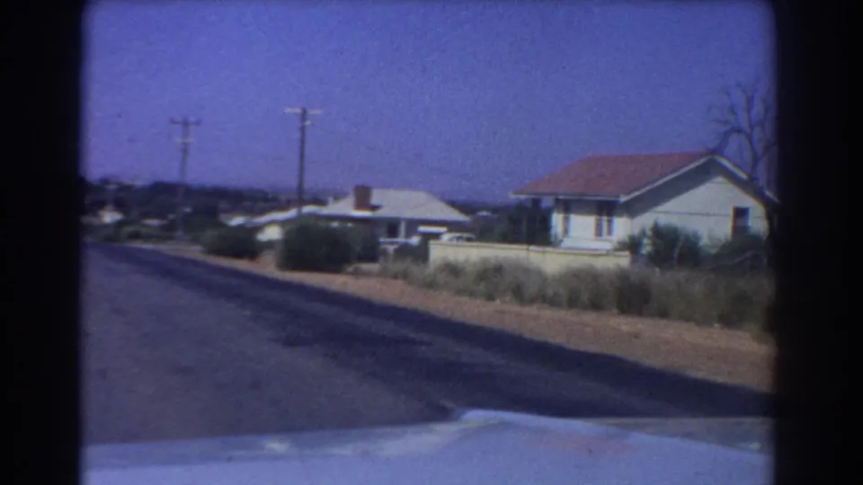 1967 Perth home-movie view along a suburban road, with a car hood at bottom, tiled-roof houses, low shrubs, and utility poles.