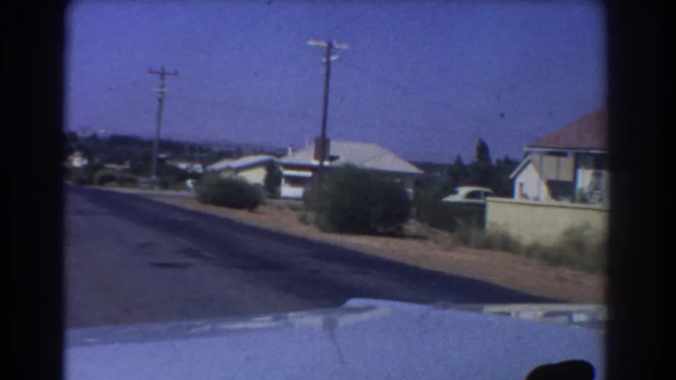 Moving 8mm view from a car in Perth, Australia, showing low suburban houses, dry roadside planting, utility poles, and part of the car hood.