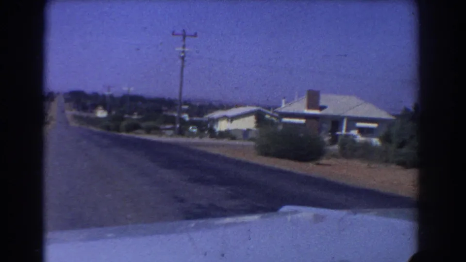 Perth suburban street view from a moving car, with a modest house, roadside shrubs, utility pole, and soft 8mm color.