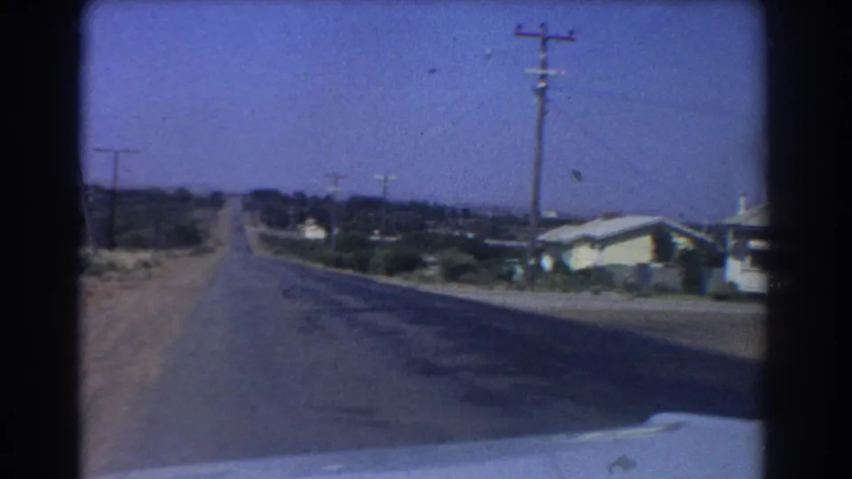 Forward-looking 1967 Perth driving view down a quiet suburban road, with houses to the side, utility poles, open verge, and car hood visible.
