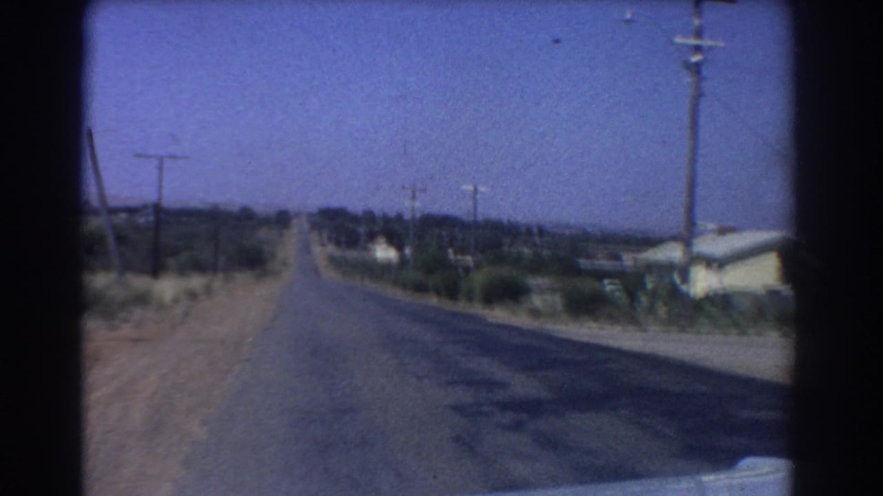 1967 Perth-area home-movie view down a straight paved road, with utility poles, dry roadside scrub, and modest houses along the edge.