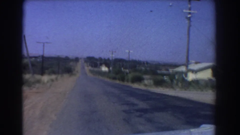 1967 Perth-area home-movie view down a straight paved road, with utility poles, dry roadside scrub, and modest houses along the edge.