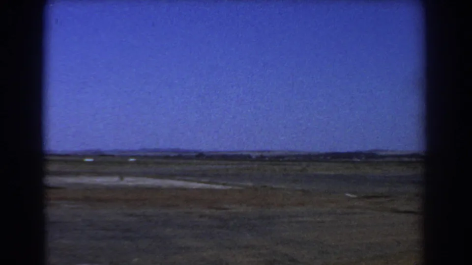 A wide Perth-area field stretches across the frame under blue sky, with sparse ground texture and the soft grain of 8mm film.