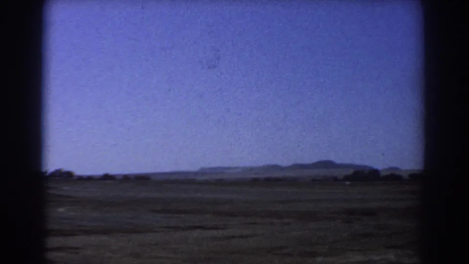 Archival 8mm frame from the Perth-area landscape shot, showing a wide field, distant low hills, and sparse trees below a pale blue sky.
