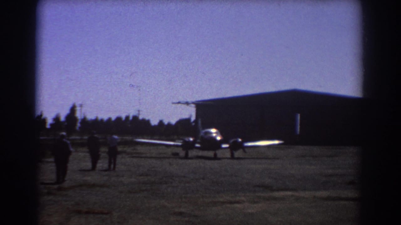 Small twin-engine aircraft on an airfield with people walking nearby and a hangar behind it.