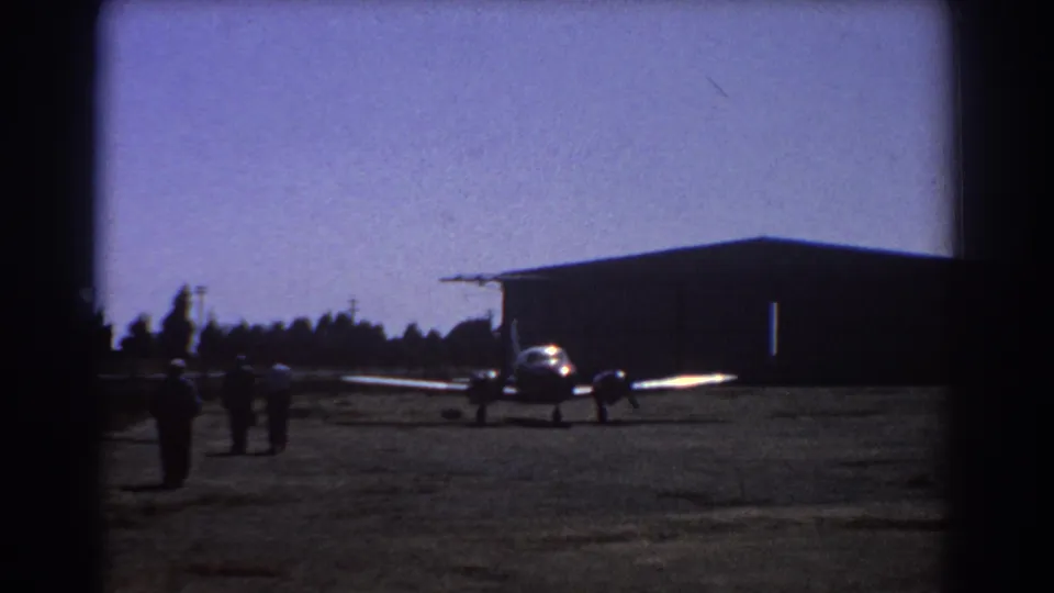 Small aircraft remains parked at a Perth airfield in 1967 while several distant figures walk across the apron near a hangar.