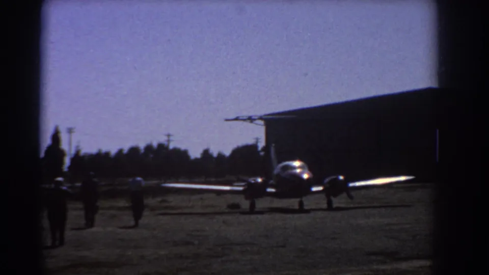 1967 Perth airfield view with distant people approaching a small twin-engine aircraft positioned beside a large hangar.