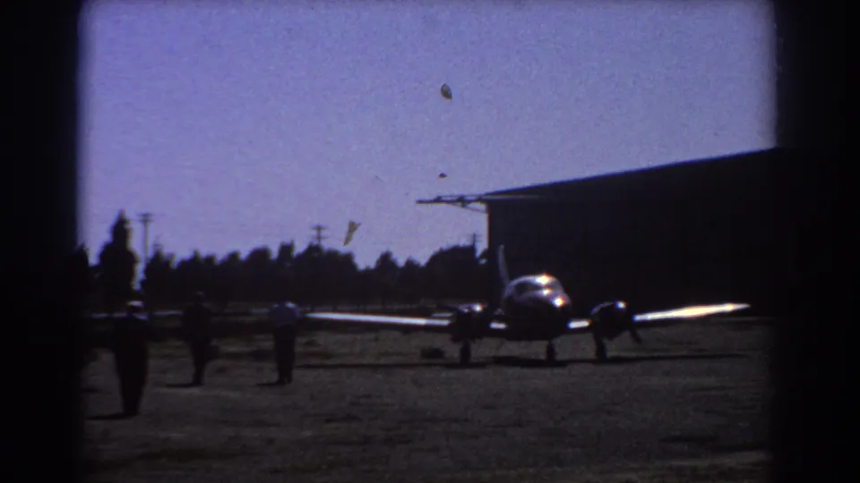 Final 1967 Perth frame shows the small twin-engine aircraft near a dark hangar, with distant people on the open airfield.