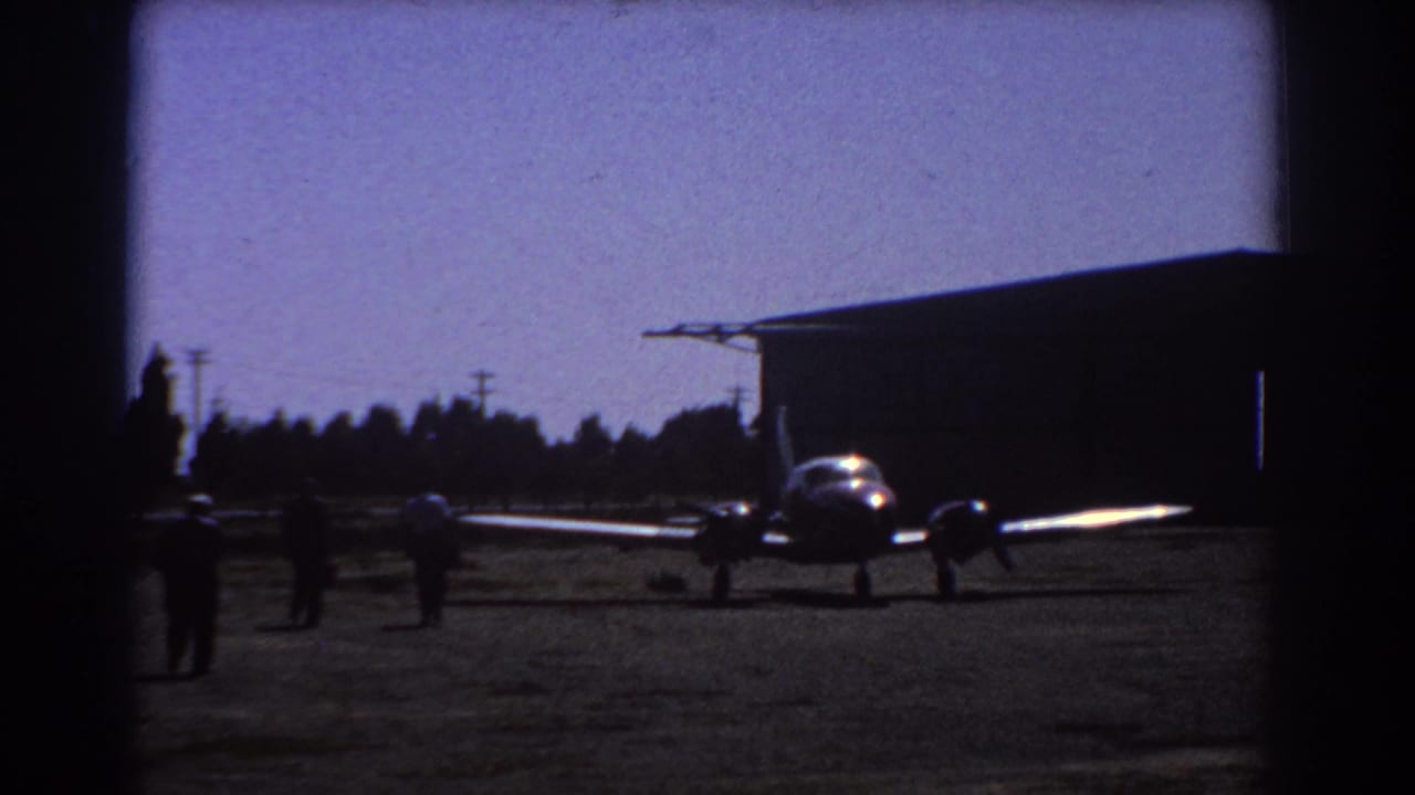 Small propeller plane parked on an airfield with people nearby and a hangar behind it.