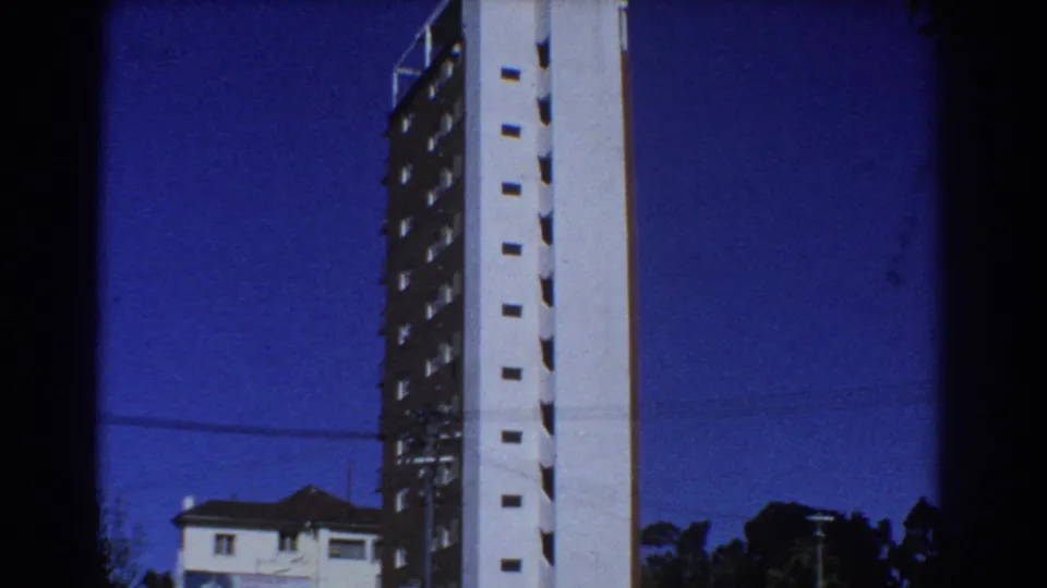 Perth home-movie frame showing a tall modern high-rise against blue sky, with a suburban roofline and trees below.
