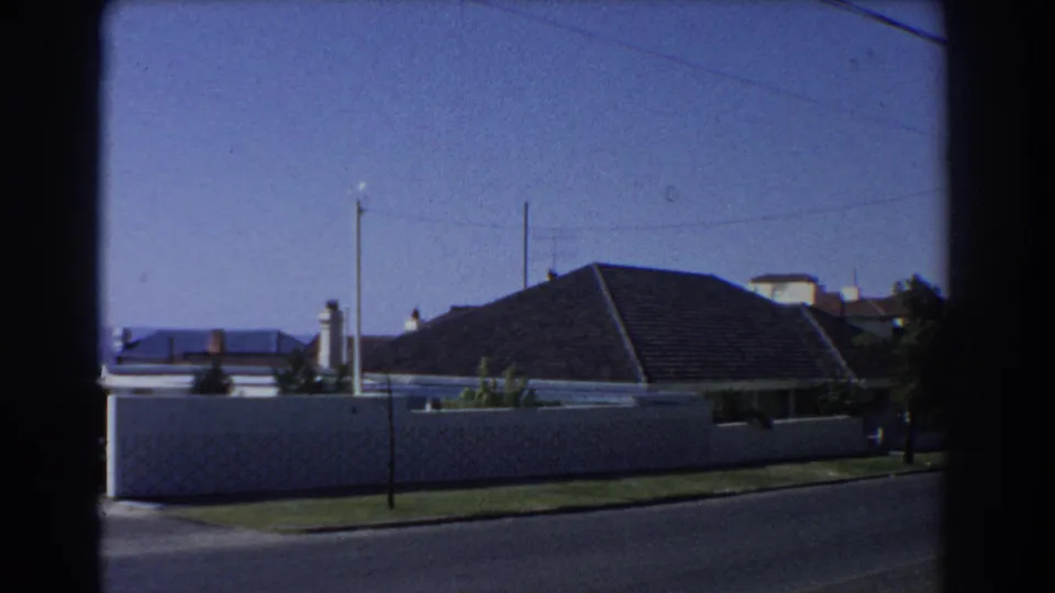 Perth 1967 home-movie frame showing a residential street, white boundary wall, tiled roofs, poles, and overhead wires.