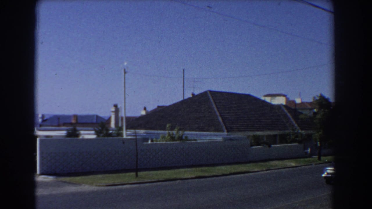 1967 Perth home-movie frame showing a quiet residential street, white boundary wall, dark tiled roof, utility poles, and blue sky.