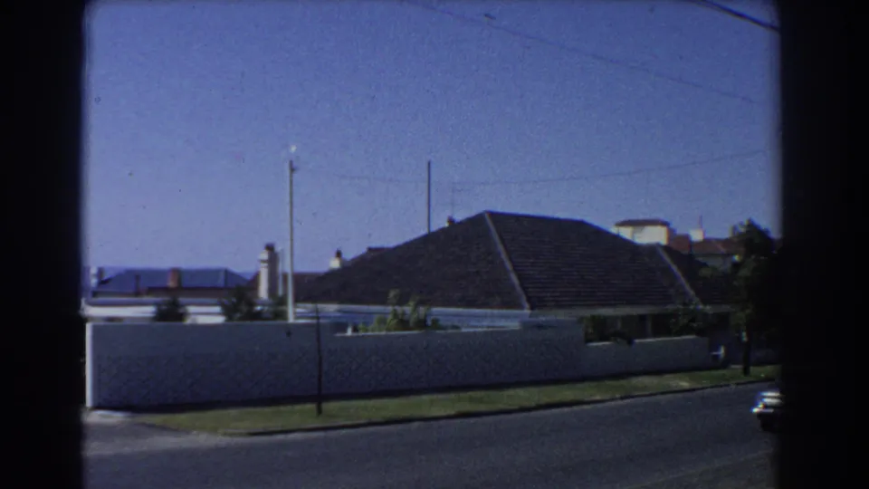 1967 Perth home-movie frame showing a quiet residential street, white boundary wall, dark tiled roof, utility poles, and blue sky.