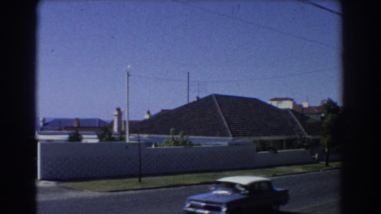 Blue and white classic car passing a white wall on a quiet suburban street.