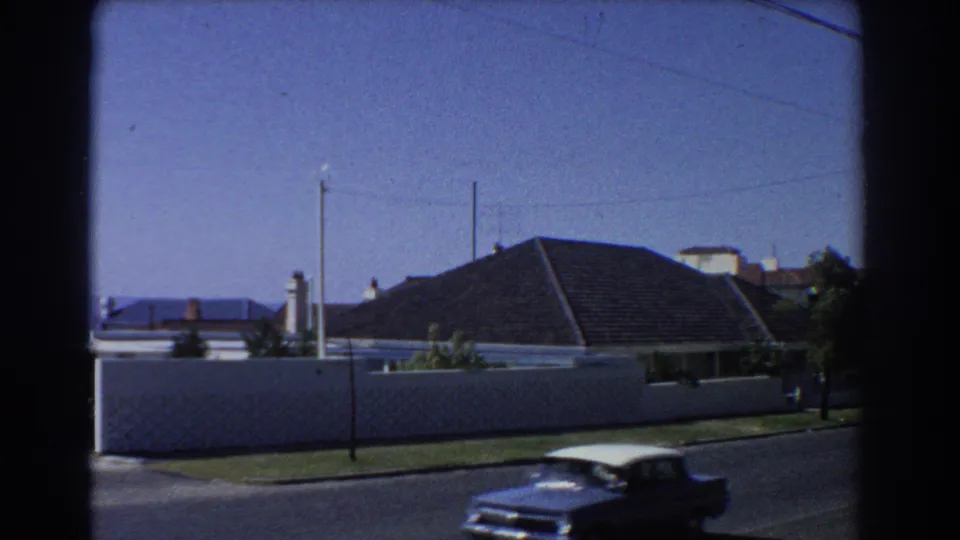 Blue and white classic car moves along a quiet Perth residential street in a 1967 8mm home-movie frame.