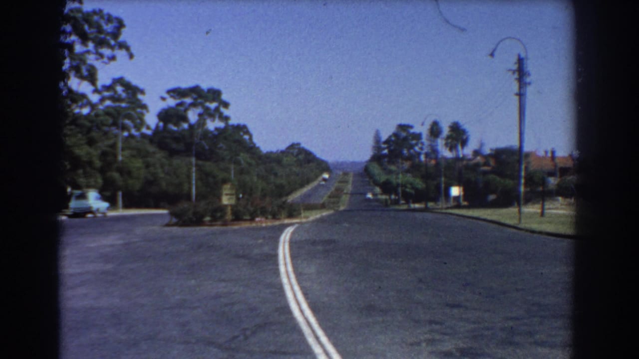 Wide suburban road curving past lawns, trees, a streetlamp, and a pale vintage car at left.