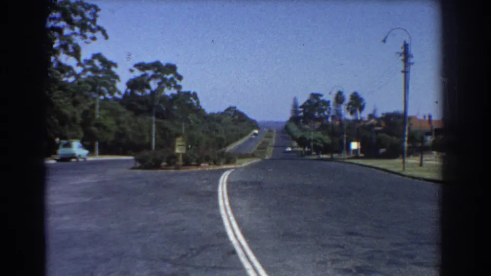 1967 Perth street view from a moving vehicle, with a broad roadway, landscaped verges, trees, a streetlamp, and distant traffic.